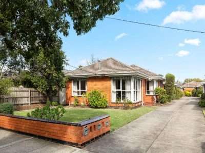 Single-story brick suburban home with a front lawn, concrete driveway, and a low brick planter under a large tree.