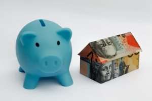 Blue piggy bank beside a house made of banknotes, illustrating savings for housing or mortgages.