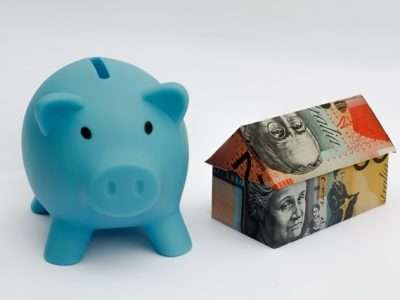Blue piggy bank beside a house made of banknotes, illustrating savings for housing or mortgages.