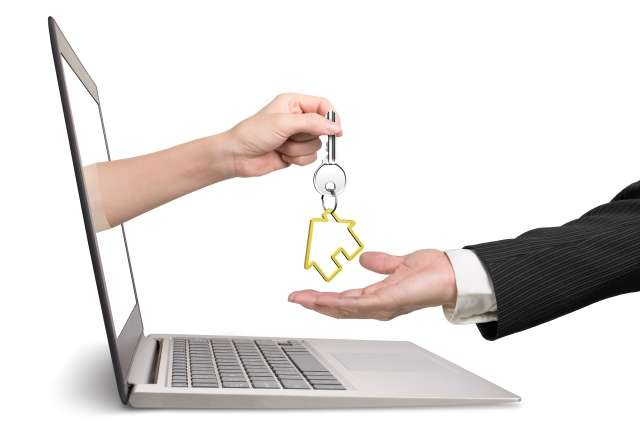Hand from a laptop handing a house key to a suited hand, symbolizing online real estate transactions.