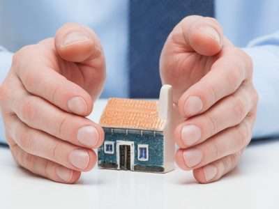 Person shielding a small model house with their hands, symbolizing home protection or insurance safety net.